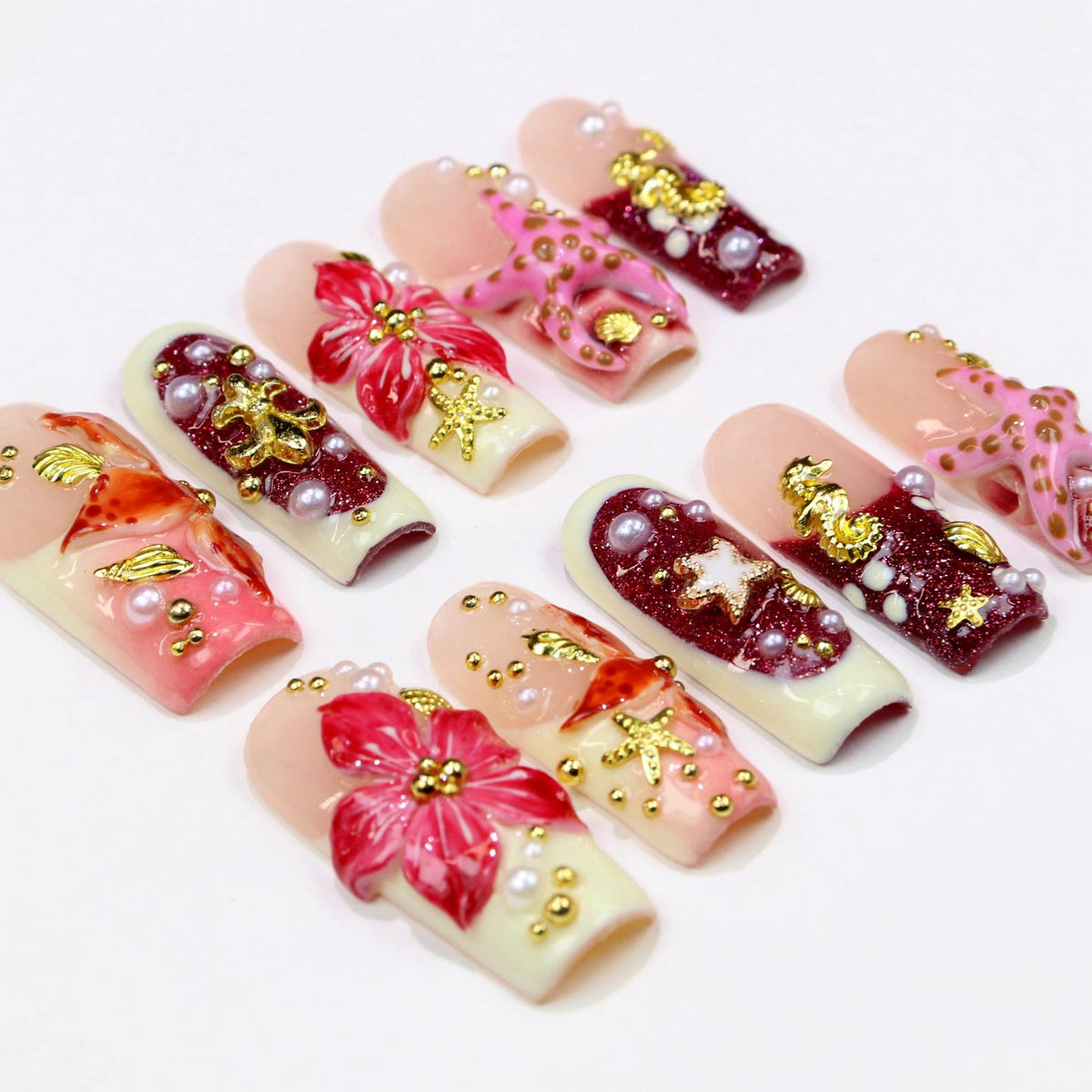 A collection of artificial nails with various designs. Some nails feature a light pink base with red and gold floral accents, while others have a dark red base with gold embellishments and pearls. The nails are arranged on a white surface.