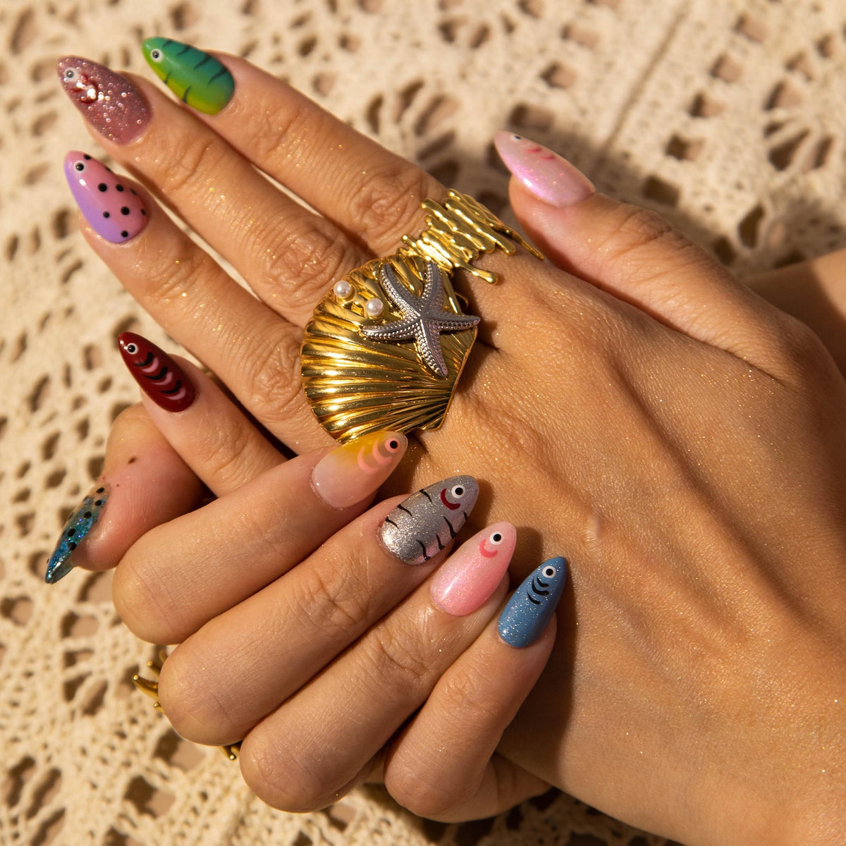 A hand with long, colorful, painted fingernails. The nails feature various designs, including fish, polka dots, and glitter. A gold ring with a seashell and starfish design is also visible.