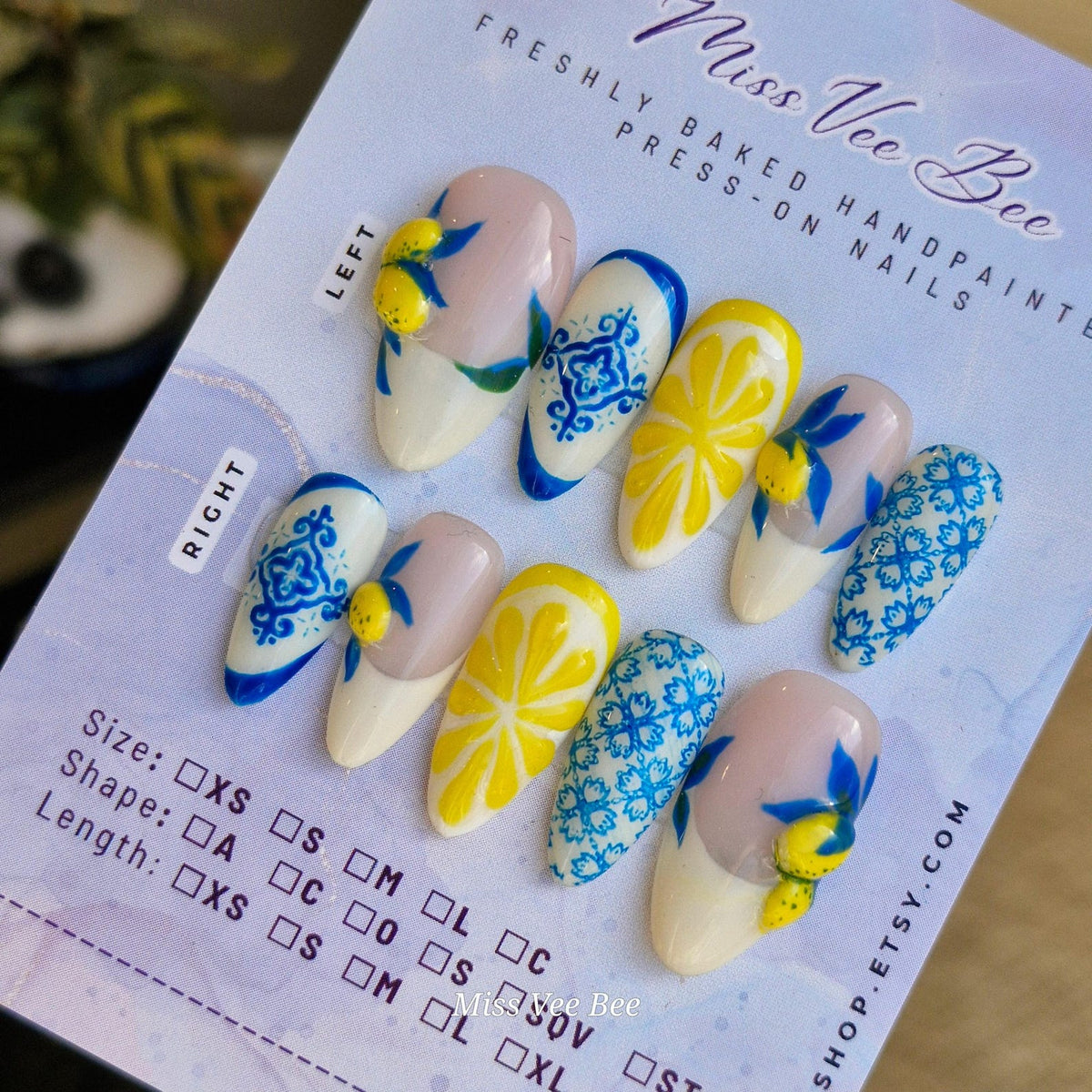 Hand-painted press-on nails with a lemon theme. The nails feature designs of lemons, blue floral patterns, and a white base. The nails are in various shapes and sizes, with a light pink and white colo