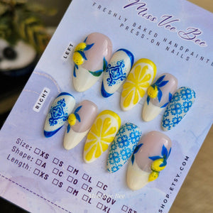 Hand-painted press-on nails with a lemon theme. The nails feature designs of lemons, blue floral patterns, and a white base. The nails are in various shapes and sizes, with a light pink and white colo
