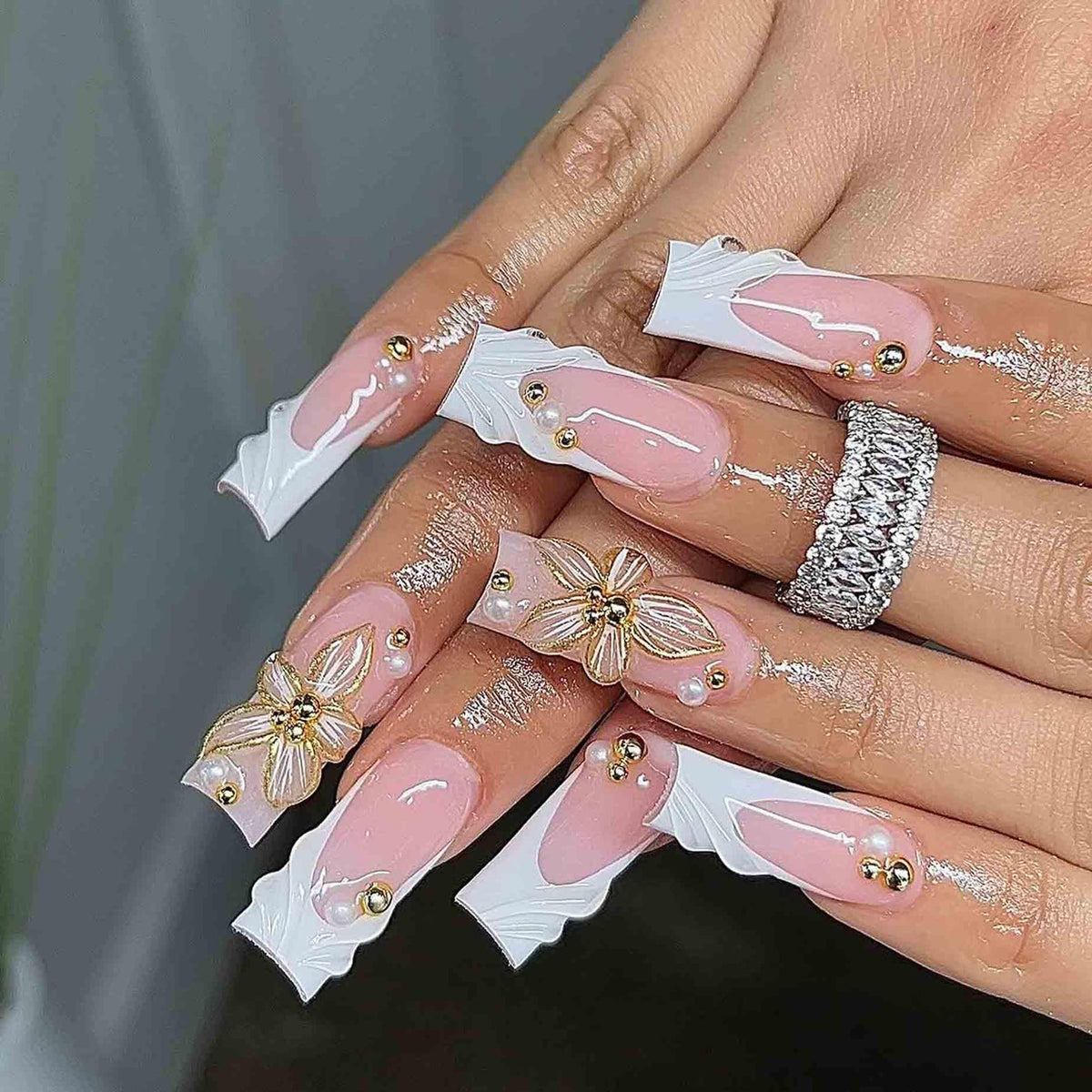 Long, square-shaped acrylic nails with a pink and white design. The nails feature a white wavy edge, pink base, gold embellishments, and 3D flower accents. A silver ring with clear stones is also visi