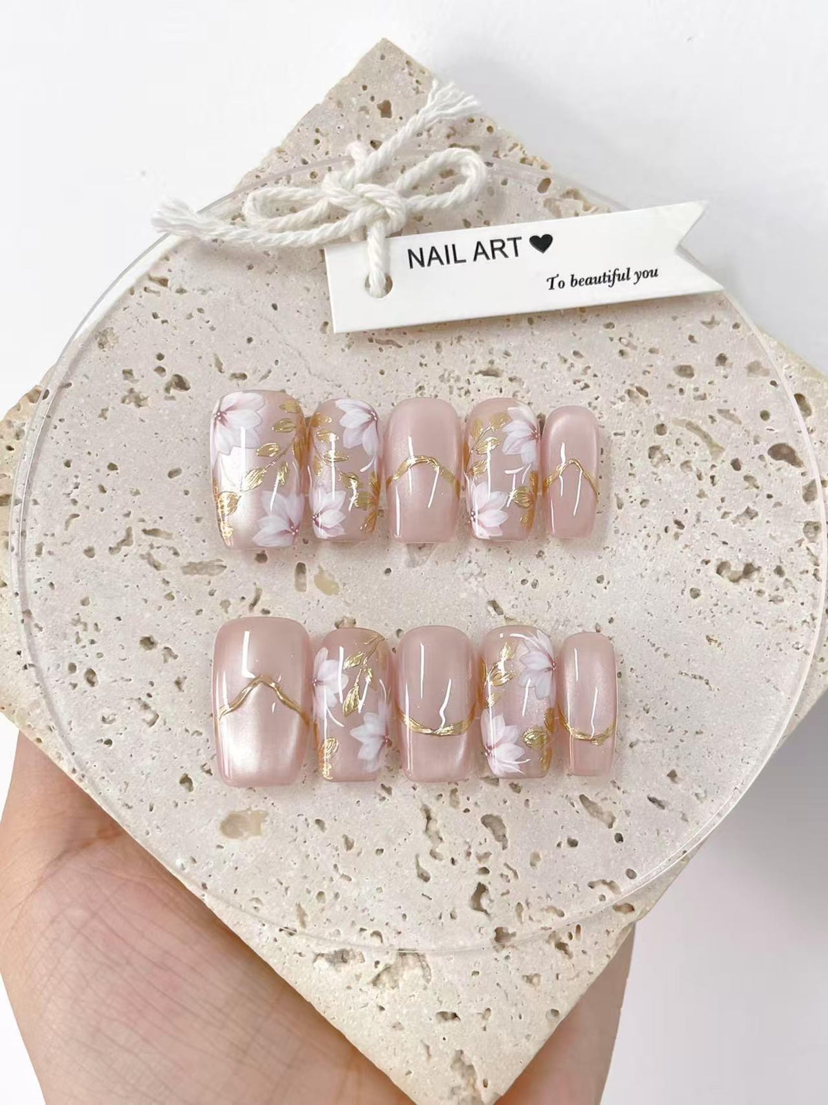 A set of artificial nails with a light pink base, adorned with white floral designs and gold accents. The nails are displayed on a textured, off-white surface, with a tag that reads "NAIL ART" and "To