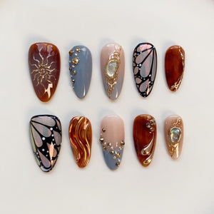 A set of ten almond-shaped, press-on nails. Designs include a sunburst, butterfly, and abstract gold accents. Colors range from brown, gray, and nude to iridescent and gold, with gem embellishments.