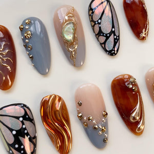 A collection of almond-shaped artificial nails in various designs. Some nails feature gold embellishments, iridescent butterfly patterns, and jewel accents. Colors include brown, gray, nude, and a shimmering orange.