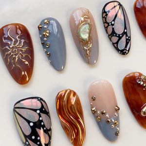 Close-up of a collection of artificial nails with various designs. The nails feature a range of colors, including brown, gray, and nude, and are adorned with gold embellishments, iridescent butterfly patterns, and textured designs.