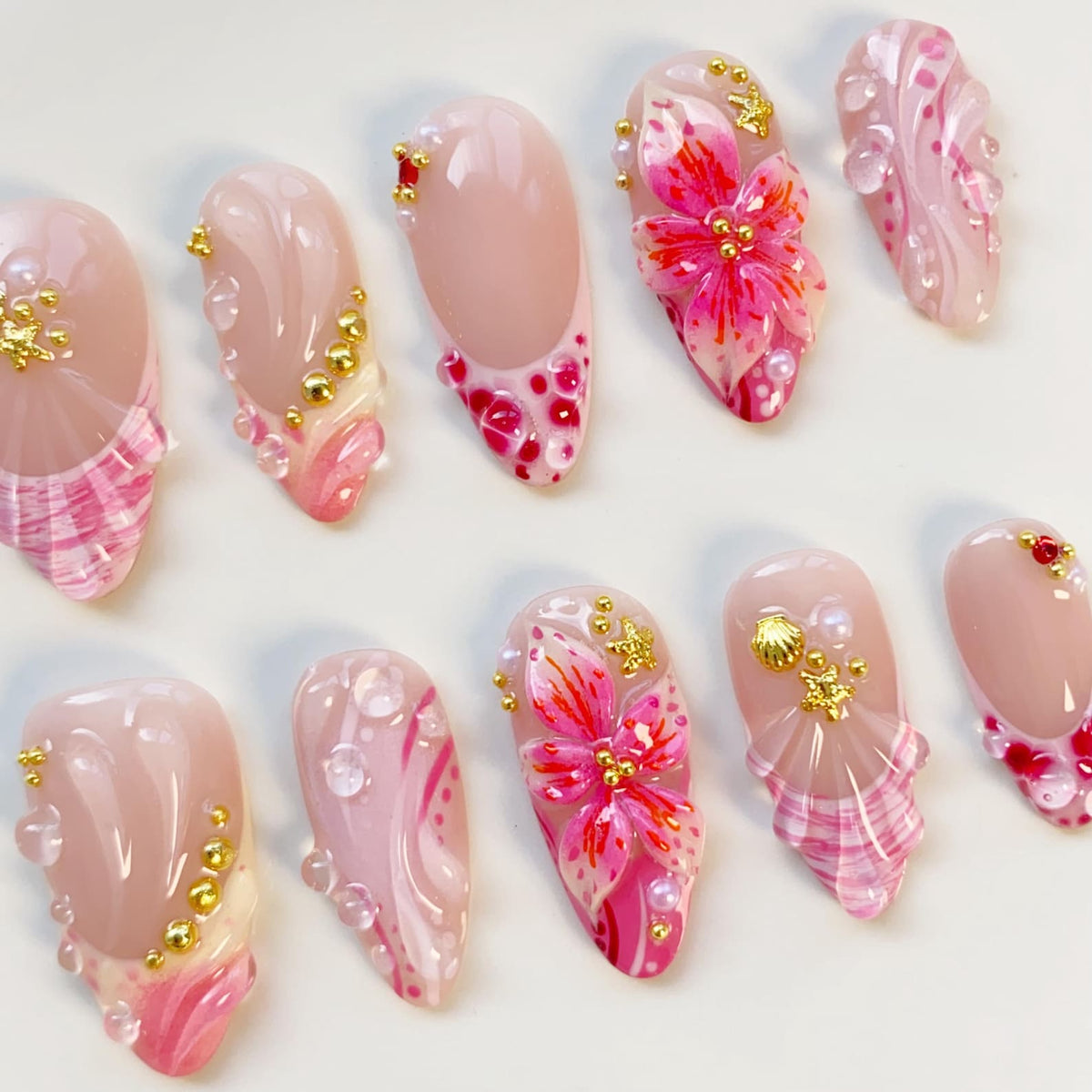A collection of ten artificial nails with various designs. The nails feature a light pink base with intricate designs, including floral patterns, gold accents, and pearl embellishments. Some nails have a clear, glossy finish, while others showcase a 