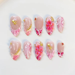 A set of ten artificial nails with a variety of designs. The nails feature a nude base with pink and white accents, floral patterns, and gold embellishments. Some nails have a plaid design at the base.