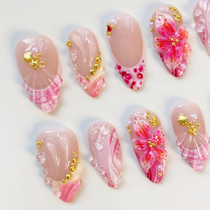 A collection of almond-shaped artificial nails. The nails feature a variety of designs, including pink and white patterns, gold embellishments, and 3D floral accents. Some nails have pearl and water droplet details.