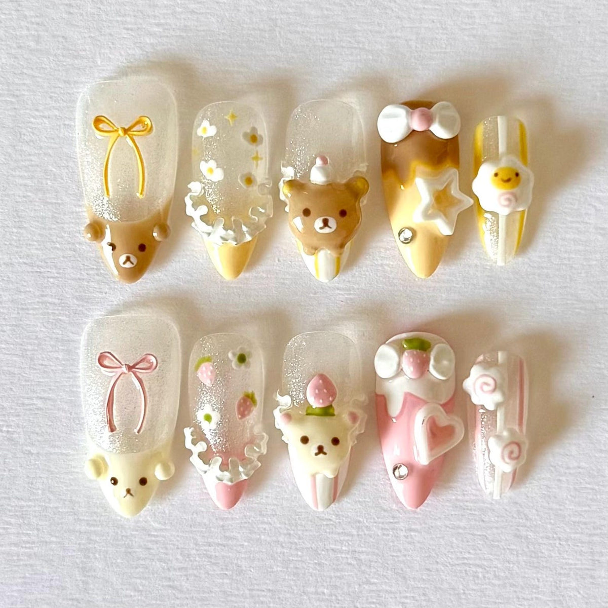 A set of ten artificial nails with various designs. The nails feature clear bases with 3D embellishments, including teddy bears, bows, flowers, and strawberries. Colors include brown, yellow, pink, and white, with glitter accents.
