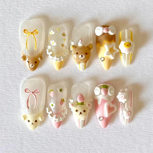 A set of ten artificial nails with various designs. The nails feature clear bases with 3D embellishments, including teddy bears, bows, flowers, and strawberries. Colors include brown, yellow, pink, and white, with glitter accents.
