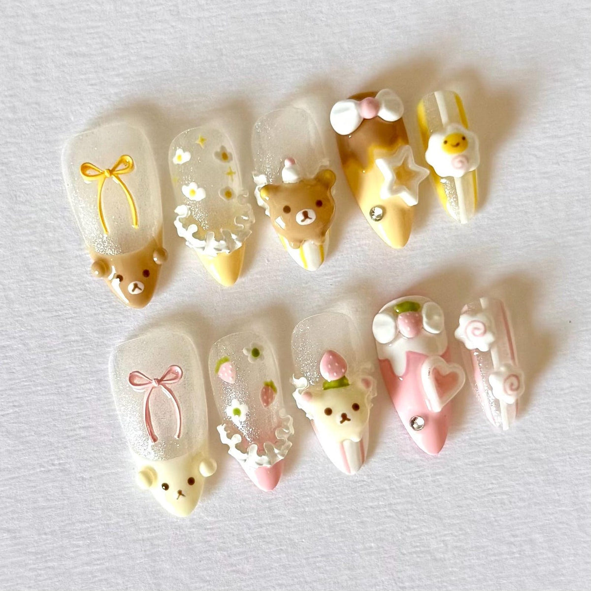 A set of ten artificial nails with various designs. The nails feature cartoon bears, bows, flowers, and stars in shades of brown, yellow, pink, and white. Some nails have glitter accents. The nails are designed for a cute and playful aesthetic.