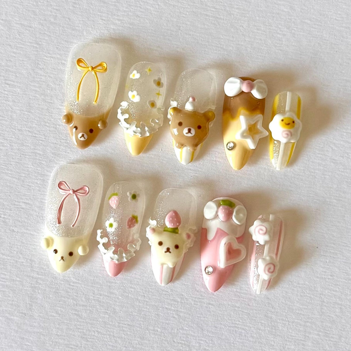 A set of ten artificial nails with various designs. The nails feature cartoon bears, bows, flowers, and other decorative elements. Colors include brown, yellow, pink, and white. Some nails have glitter accents and 3D embellishments.