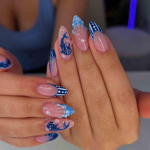 A hand with long, almond-shaped nails featuring a blue and white nautical design. The nails have a clear base with blue tips, polka dots, and whale illustrations. Some nails have a French tip design.