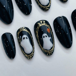 A set of press-on nails featuring a glossy black base with white speckles. Two nails display a white ghost illustration framed in gold, with one ghost holding a small orange balloon. The nails have an