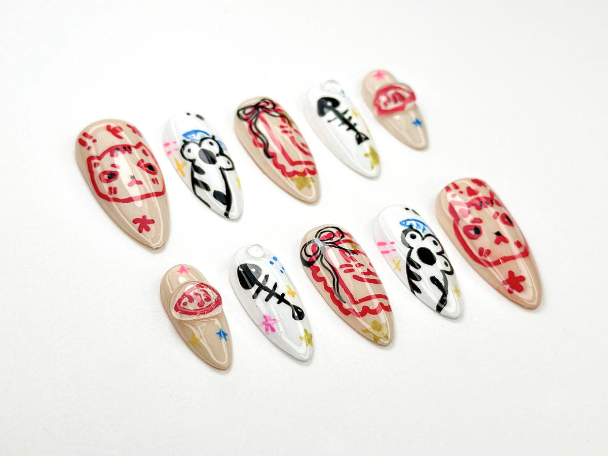 A collection of ten almond-shaped false nails, each hand-painted with unique designs. The nails feature cartoon cat faces, fish bones, and abstract patterns in red, black, and white on a neutral beige base. Some nails have star and flower accents.