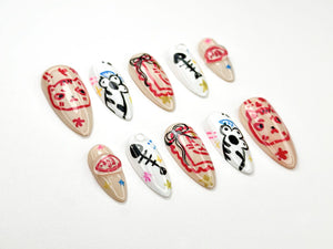 A collection of ten almond-shaped false nails, each hand-painted with unique designs. The nails feature cartoon cat faces, fish bones, and abstract patterns in red, black, and white on a neutral beige base. Some nails have star and flower accents.