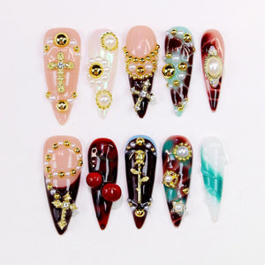 A collection of ten artificial nails, each with a unique design. The nails feature various colors, including pink, red, and teal, and are adorned with gold-colored embellishments, pearls, and crosses. Some nails have a marbled effect.