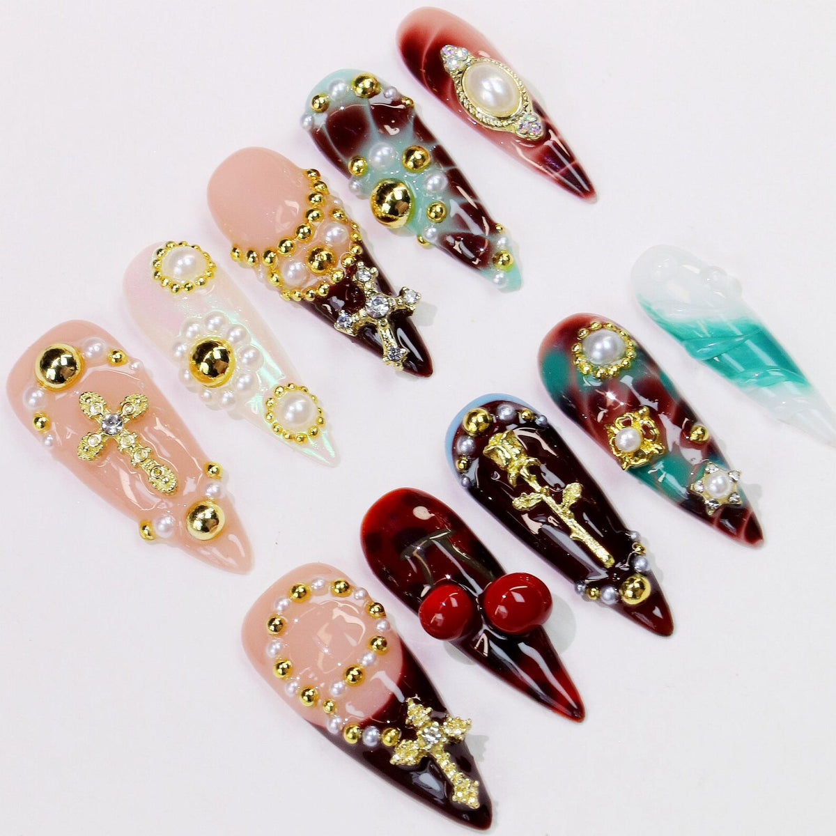 A collection of long, pointed artificial nails in various designs. Some nails feature pearl and gold accents, crosses, and other decorative elements. Colors include red, pink, and teal, with a glossy finish.
