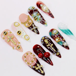 A collection of long, pointed artificial nails in various designs. Some nails feature pearl and gold accents, crosses, and other decorative elements. Colors include red, pink, and teal, with a glossy finish.