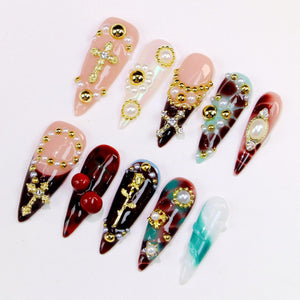 A collection of ten artificial nails in various designs. The nails feature a range of colors, including pink, red, and teal, and are adorned with gold crosses, pearls, and other decorative elements. Some nails have a glossy finish, while others have 