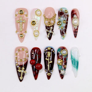 A collection of ten long, pointed artificial nails. Each nail features a unique design with embellishments like gold crosses, pearls, and decorative accents. The nails are painted in various colors, including pink, red, and teal.