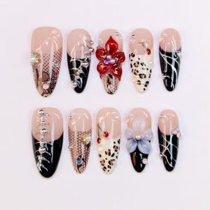 A collection of ten artificial nails with various designs. Some nails feature black tips, lace patterns, and silver embellishments. Others showcase floral accents, leopard print, and pearl-like beads. The nails have a glossy finish and a neutral base