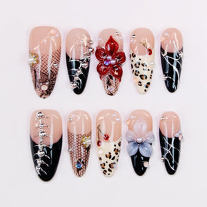 A set of ten artificial nails with various designs. The nails feature a nude base with black, white, and red accents. Some nails have floral embellishments, while others showcase patterns, rhinestones, and pearls.