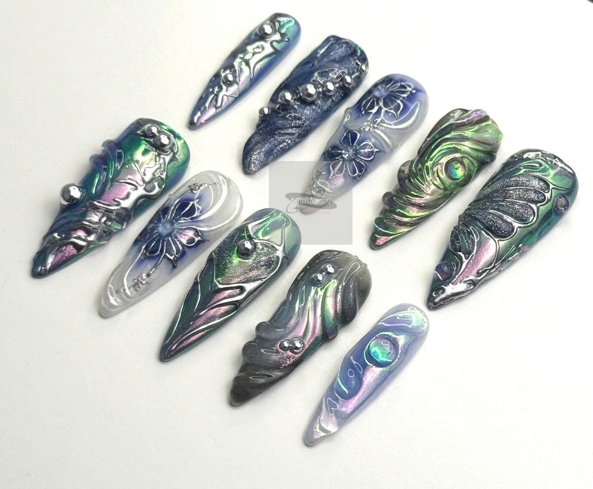 A collection of eleven long, pointed artificial nails. The nails feature a variety of designs, including floral patterns, metallic accents, and iridescent finishes in shades of blue, green, and silver