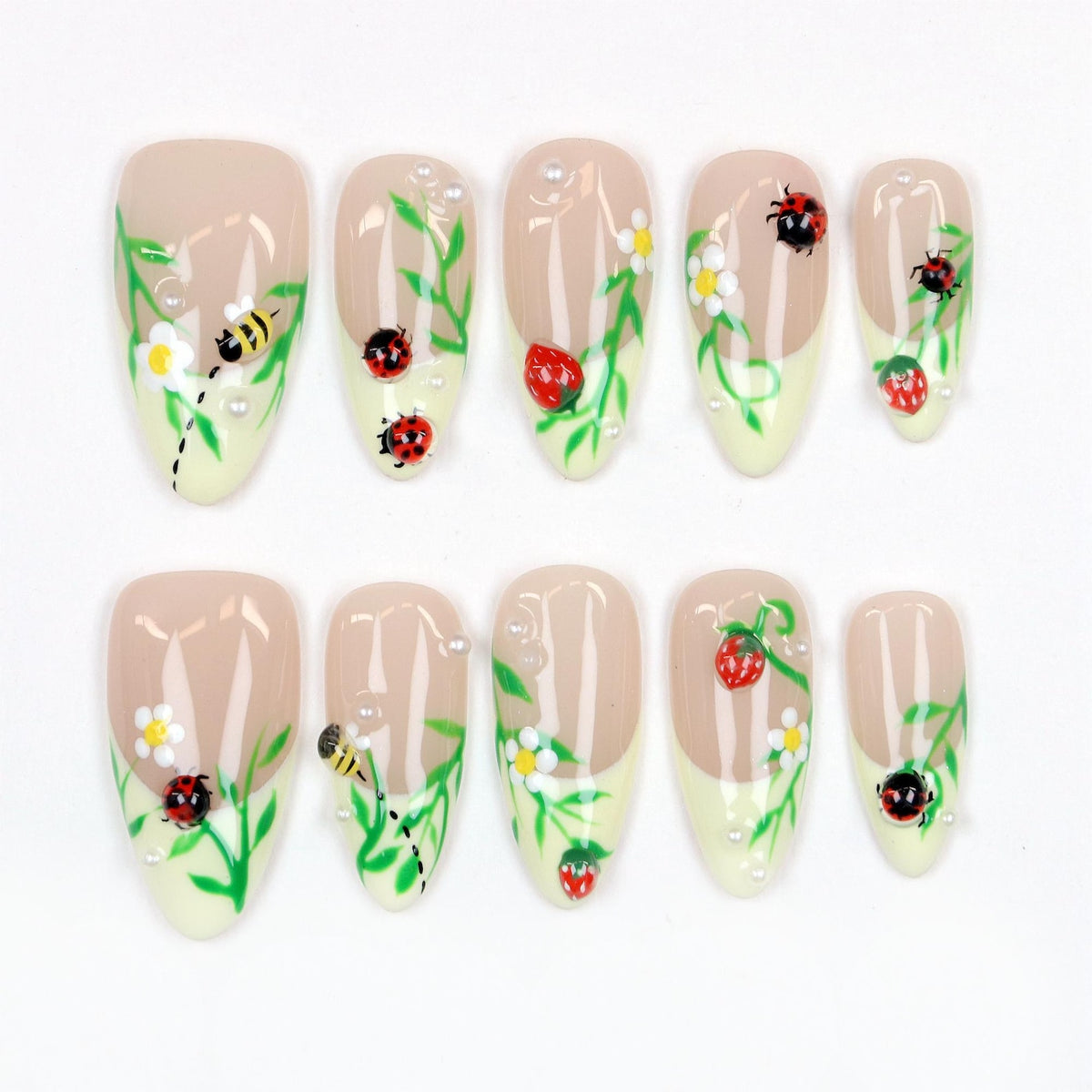 A set of ten almond-shaped false nails. Each nail features a light beige base with a yellow-green tip. The nails are decorated with ladybugs, strawberries, bees, white flowers, green vines, and small pearl accents.