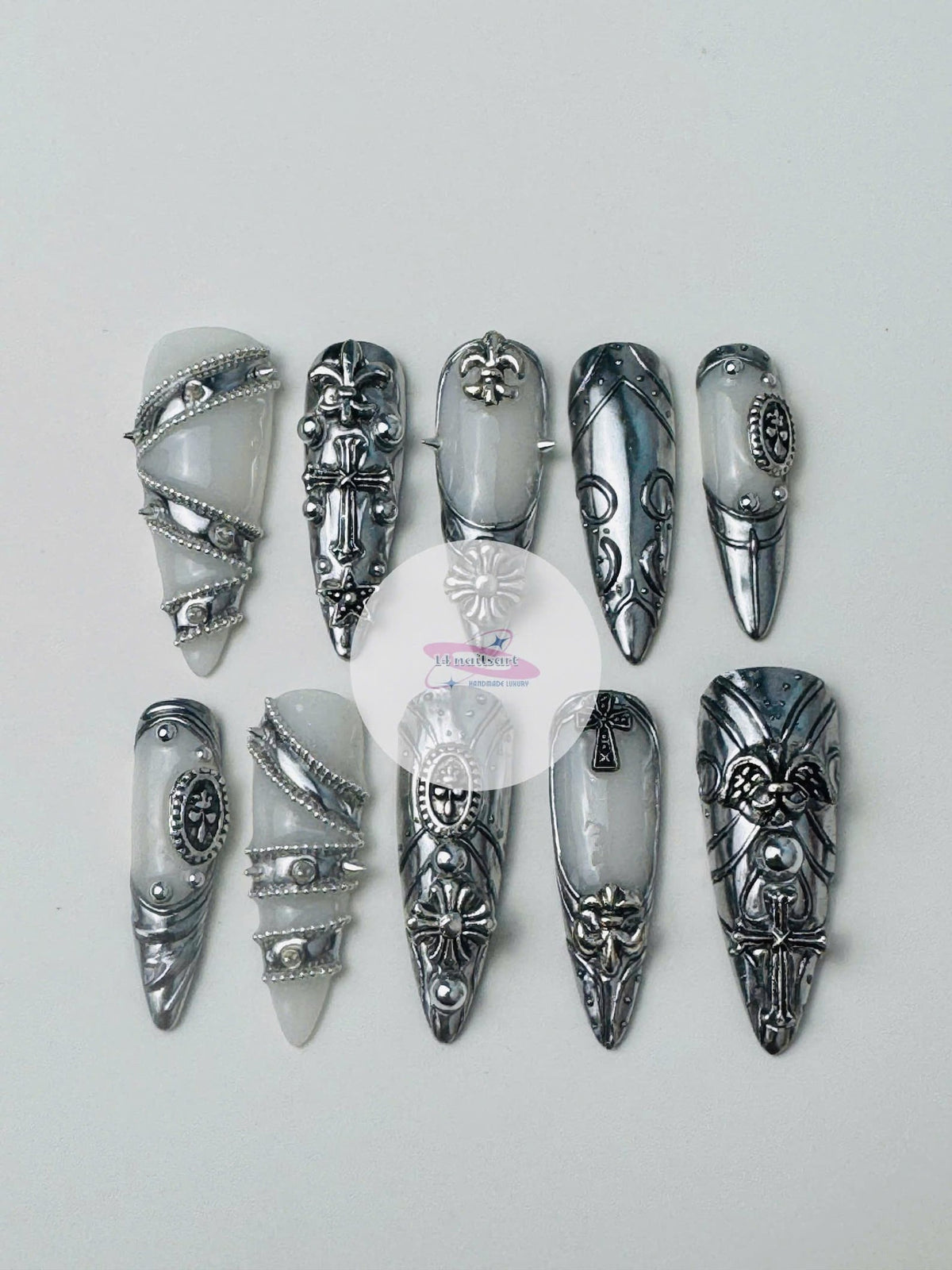 A set of ten long, silver and white press-on nails. Each nail features intricate silver embellishments, including crosses, fleur-de-lis, and ornate designs. The nails have a glossy finish and a pointe