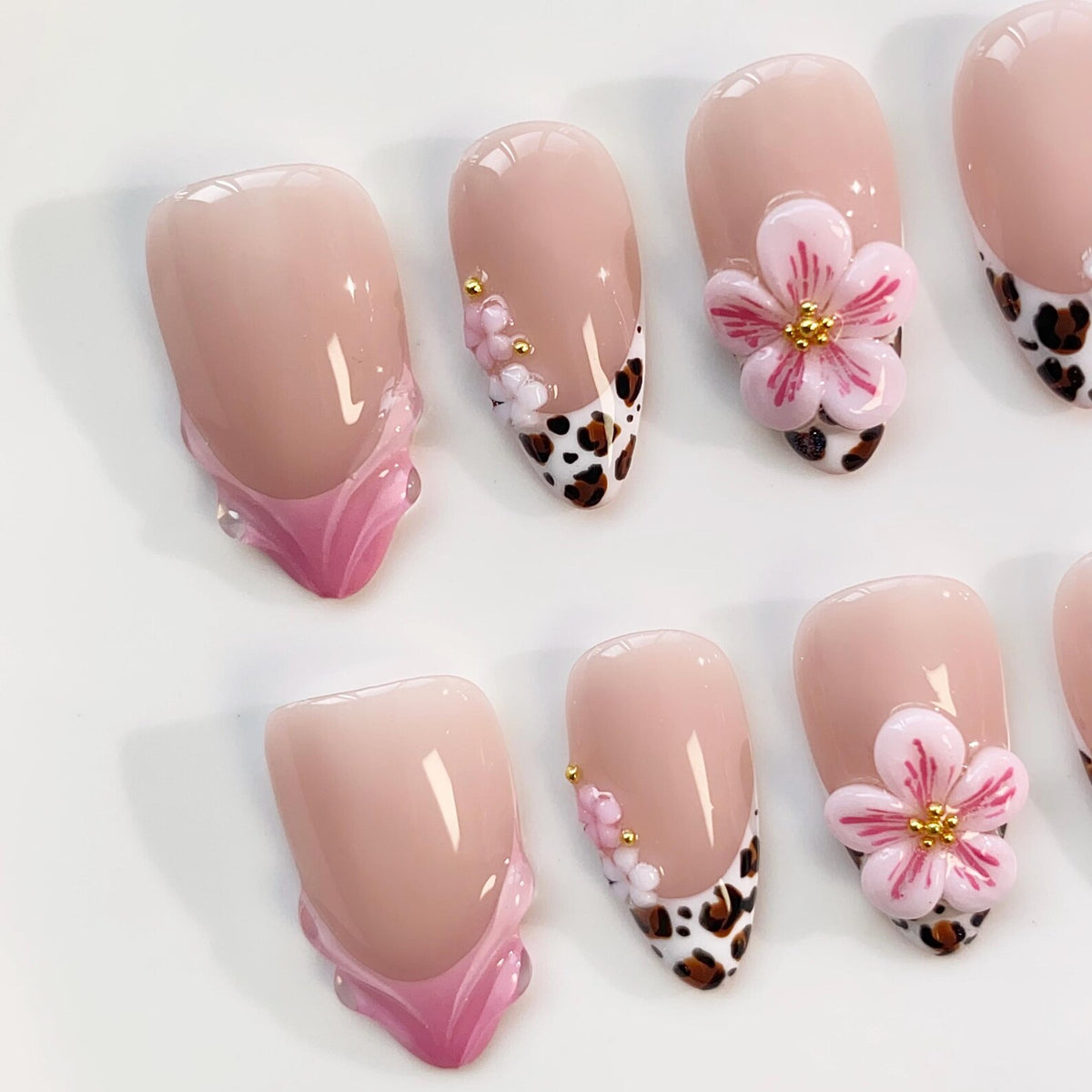 A set of almond-shaped artificial nails. The nails feature a light pink base with various designs, including a French tip with a leopard print, small gold accents, and 3D pink flower embellishments. The nails are arranged on a white surface.