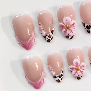 A set of almond-shaped artificial nails. The nails feature a light pink base with various designs, including a French tip with a leopard print, small gold accents, and 3D pink flower embellishments. The nails are arranged on a white surface.