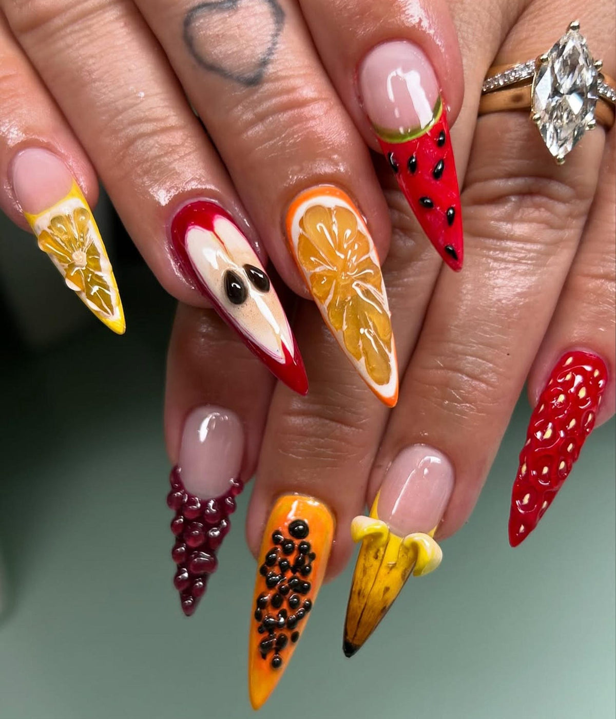 A set of long, pointed fingernails with detailed fruit designs. Each nail features a different fruit, including a lemon, apple, orange slice, watermelon, grapes, papaya, banana, and strawberries. The nails are painted with vibrant colors and 3D textu