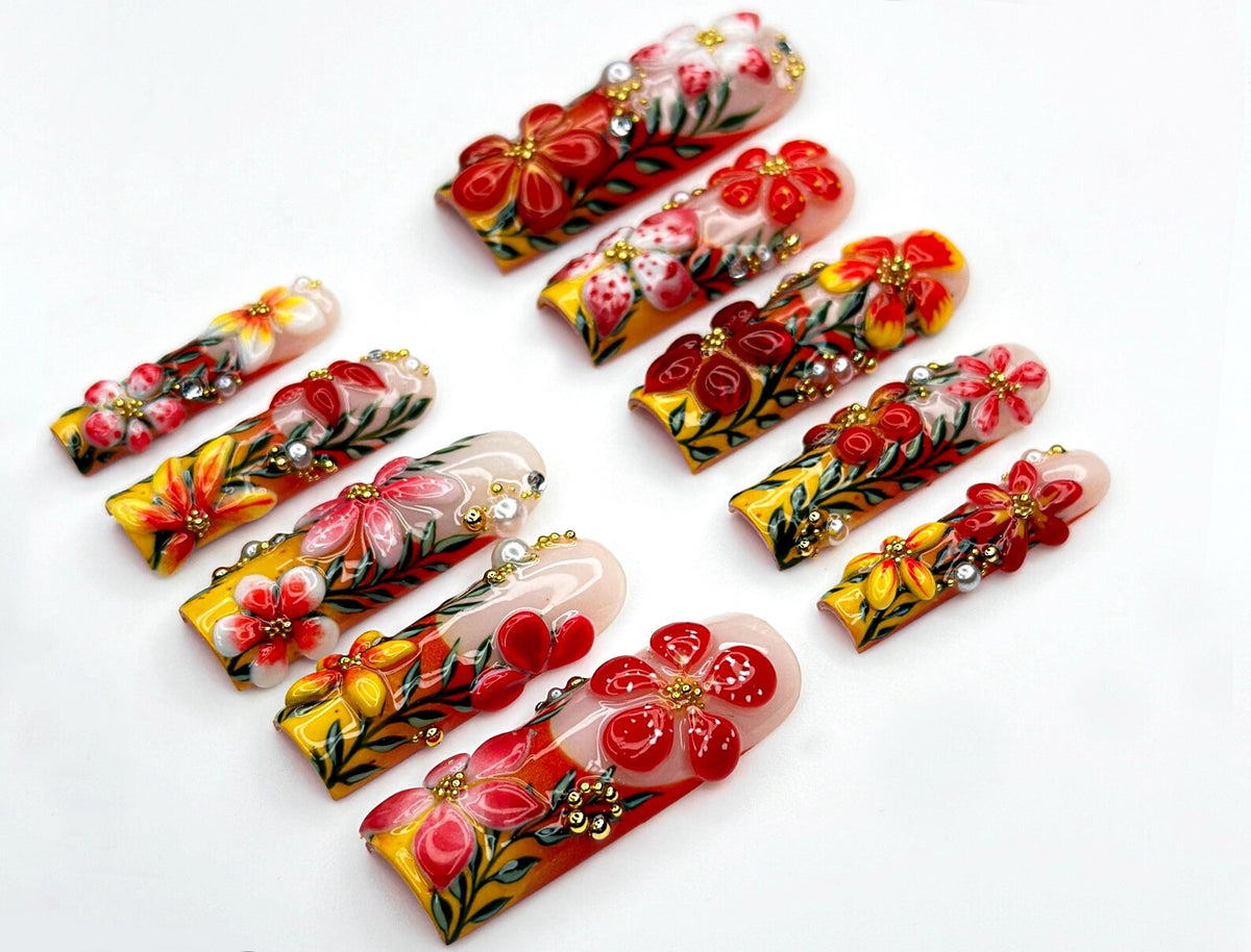 A collection of press-on nails with intricate floral designs. Each nail features a base of yellow, red, and white, adorned with detailed flower art, gold accents, and small embellishments. The nails a
