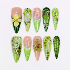 A collection of ten artificial nails with various designs. The nails feature a range of colors, including green, pink, and gold, with floral and abstract patterns. Some nails have 3D embellishments, while others have glitter and pearl accents.