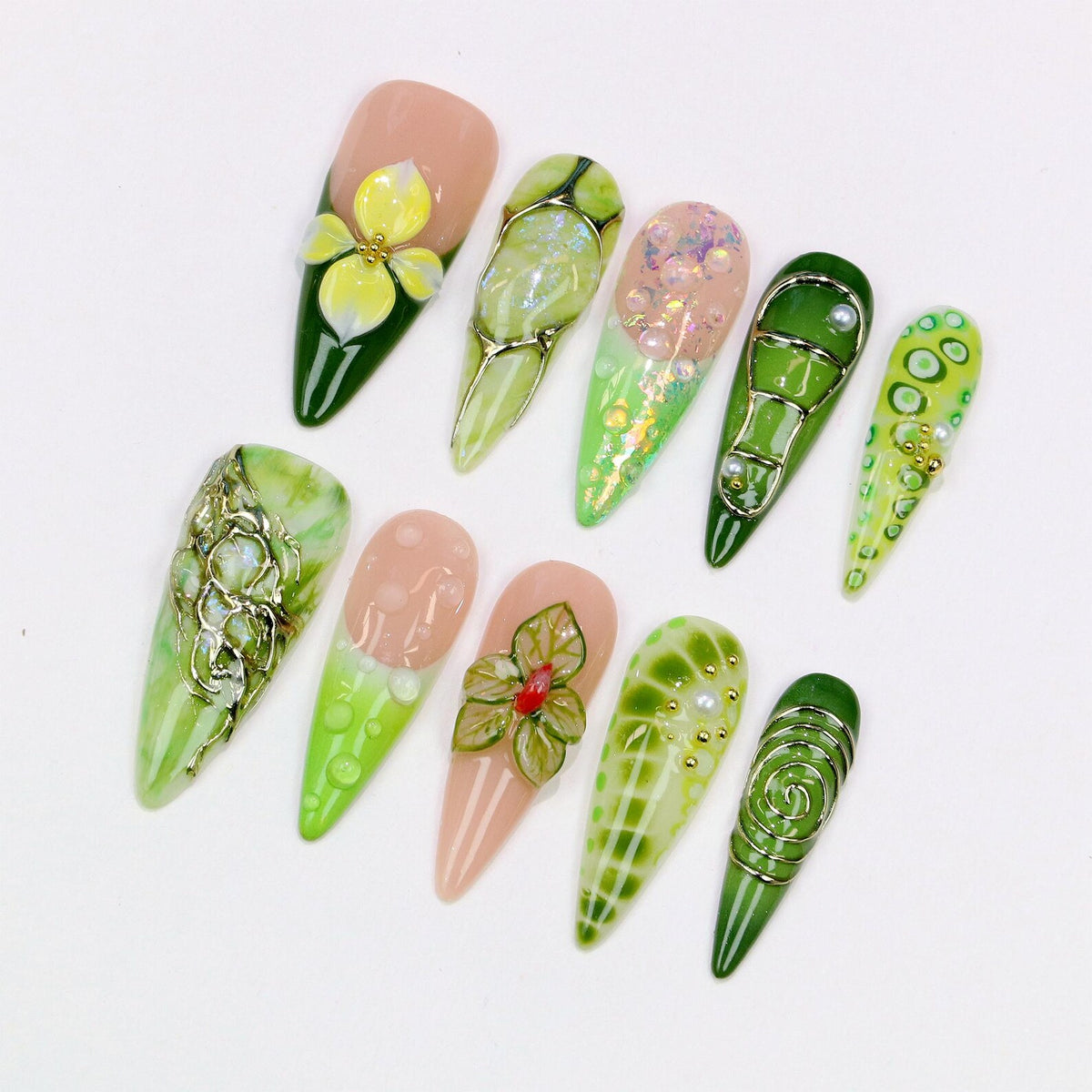 A collection of ten almond-shaped artificial nails with various designs. The nails feature a range of colors, including green, pink, and gold, with floral and abstract patterns. Some nails have 3D embellishments, such as flowers and gems.