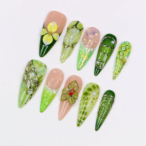 A collection of ten almond-shaped artificial nails with various designs. The nails feature a range of colors, including green, pink, and gold, with floral and abstract patterns. Some nails have 3D embellishments, such as flowers and gems.