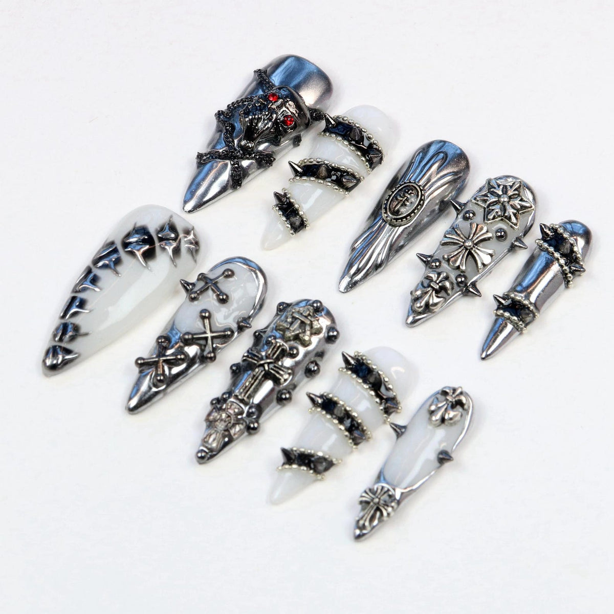 A collection of silver-toned nail art designs. The designs include ornate metal accents, crosses, spikes, and skull details. Some nails are white with silver embellishments, while others are entirely silver.
