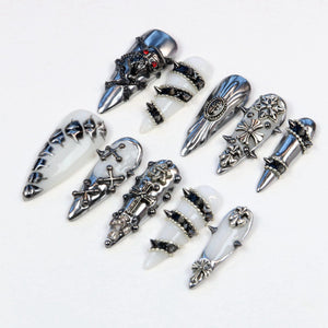 A collection of silver-toned nail art designs. The designs include ornate metal accents, crosses, spikes, and skull details. Some nails are white with silver embellishments, while others are entirely silver.
