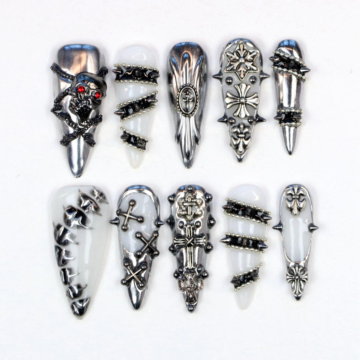 A collection of ten long, pointed nail designs. The nails feature silver and black embellishments, including skulls, crosses, spikes, and geometric patterns. Some nails have a white base, while others are silver.
