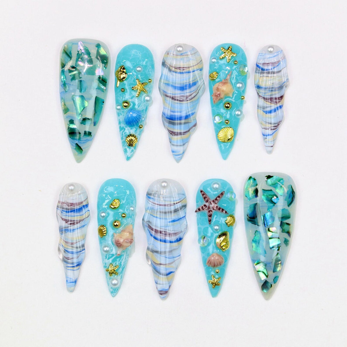 A set of ten artificial nails with a beach theme. The nails are various shapes and sizes, with a light blue base and decorated with seashells, starfish, and iridescent accents. Some nails have a swirl pattern.
