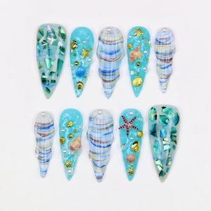 A set of ten artificial nails with a beach theme. The nails are various shapes and sizes, with a light blue base and decorated with seashells, starfish, and iridescent accents. Some nails have a swirl pattern.