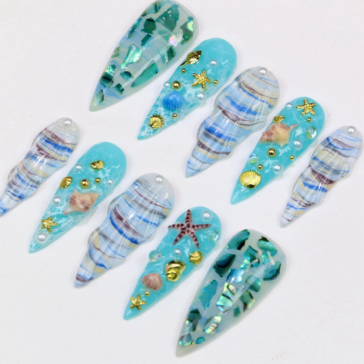 A collection of ten long, pointed artificial nails with a beach theme. The nails feature a variety of designs, including blue and white swirls, seashells, starfish, and gold accents. Some nails have a clear, glossy finish, while others have a texture