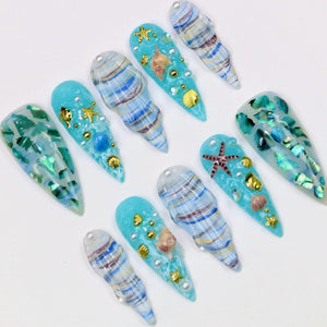 A collection of ten long, pointed artificial nails. Each nail features a light blue base with various embellishments, including seashells, starfish, gold accents, and iridescent flakes. Some nails have a swirl pattern.