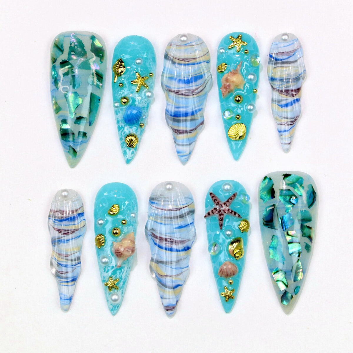 A set of ten long, pointed artificial nails. The nails feature a variety of designs, including turquoise and blue swirls, iridescent shell fragments, and gold seashell and starfish embellishments. Some nails have pearl accents.