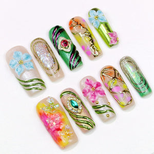An assortment of artificial nails, each with unique designs. The nails feature various colors, including pink, green, and yellow, and are adorned with floral and jewel embellishments. Some nails have gold wire accents.