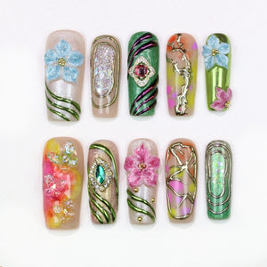 A collection of ten artificial nails with various designs. The nails feature floral embellishments, glitter, and metallic accents. Colors include blue, pink, green, and gold. Some nails have a glossy finish, while others have a matte appearance.