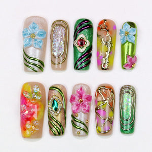 A collection of ten artificial nails with various designs. The nails feature a range of colors, including green, pink, gold, and blue, with embellishments such as flowers, glitter, and gemstones. The designs are intricate and artistic, showcasing nai