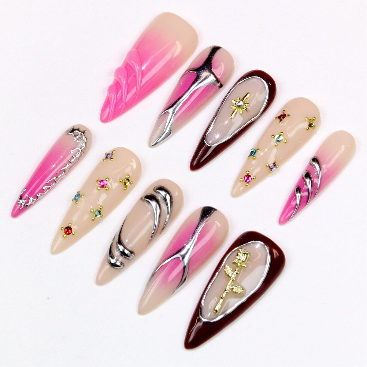A collection of ten long, pointed artificial nails. The nails feature various designs, including pink and nude gradients, silver and gold embellishments, and jewel accents. The nails are arranged on a white surface.