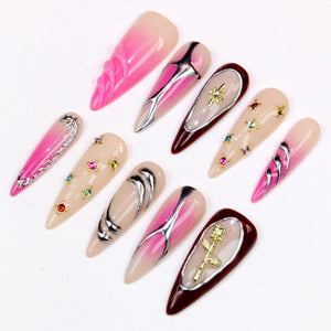 A collection of ten long, pointed artificial nails. The nails feature various designs, including pink and nude gradients, silver and gold embellishments, and jewel accents. The nails are arranged on a white surface.
